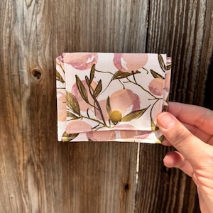 May include: A small, folded wallet with a peach and leaf pattern. The wallet is primarily white with pink and peach-colored fruit and green leaves. It has a gold-colored button closure.