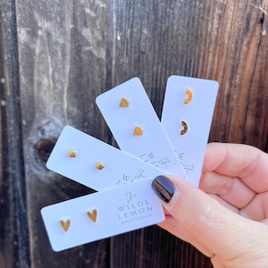 May include: Four cards with gold-colored geometric shapes on white backgrounds. The shapes are a star, a heart, a half-circle, and a triangle. The cards are held in a hand. The text on the bottom card reads "The Wilde Lemon @thewildelemon".