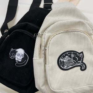 May include: Two corduroy crossbody bags with zipped pockets and embroidered patches. The black bag has a skull and cat patch, while the white bag has a cat and mountain scene patch.