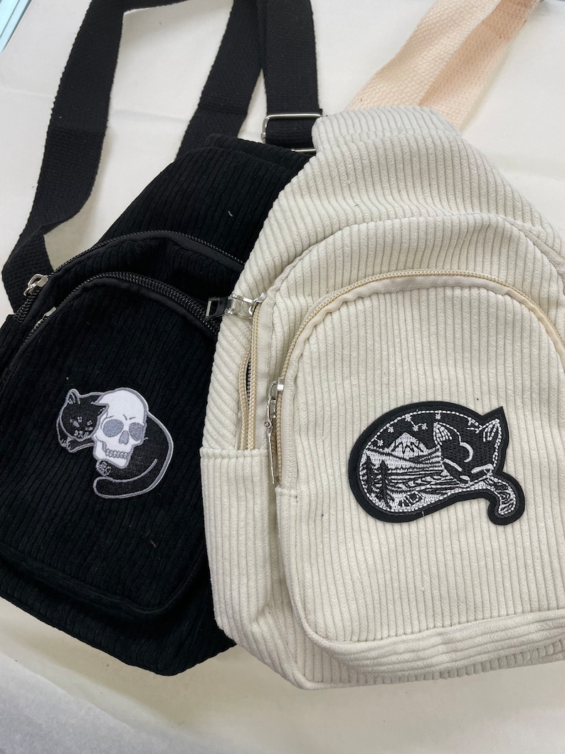 May include: Two corduroy crossbody bags with zipped pockets and embroidered patches. The black bag has a skull and cat patch, while the white bag has a cat and mountain scene patch.