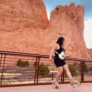 May include: A person wearing a black tank top and black shorts is roller skating on a paved path. The person is wearing a white bag with a blue design. The background is a red rock formation.