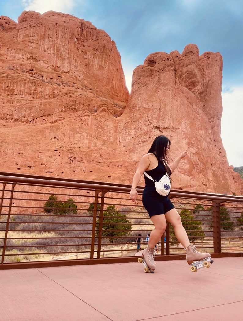 May include: A person wearing a black tank top and black shorts is roller skating on a paved path. The person is wearing a white bag with a blue design. The background is a red rock formation.