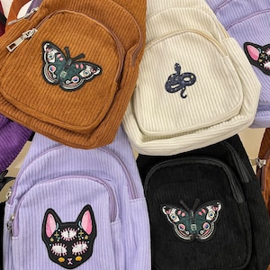 May include: Five small corduroy backpacks with different embroidered patches. The backpacks are brown, white, purple, black, and brown. The patches include a butterfly, a snake, a cat, a butterfly, and a cat.