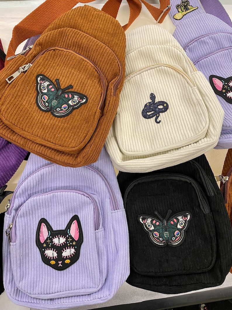May include: Five small corduroy backpacks with different embroidered patches. The backpacks are brown, white, purple, black, and brown. The patches include a butterfly, a snake, a cat, a butterfly, and a cat.