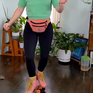 K&ouml;nnte beinhalten: Eine Person tr&auml;gt ein gr&uuml;nes T-Shirt, schwarze Leggings und eine rosa Bauchtasche und rollt auf einem Holzboden Schlittschuh. Die Person tr&auml;gt rosa Rollschuhe mit gelben Schn&uuml;rsenkeln und orangefarbenen Rollen.