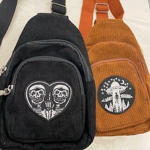 May include: Two corduroy crossbody bags. The black bag has a white embroidered patch of two skeletons in a heart. The brown bag has a white embroidered patch of an alien spaceship with the text "I want to leave".