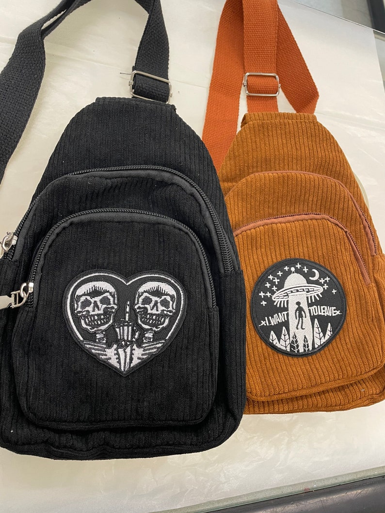 May include: Two corduroy crossbody bags. The black bag has a white embroidered patch of two skeletons in a heart. The brown bag has a white embroidered patch of an alien spaceship with the text "I want to leave".