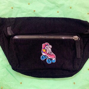K&ouml;nnte beinhalten: Schwarze Cord-Bauchtasche mit einem rosa-blauen Rollschuh-Patch. Der Patch hat einen gelben Stern und wei&szlig;e Schn&uuml;rsenkel.