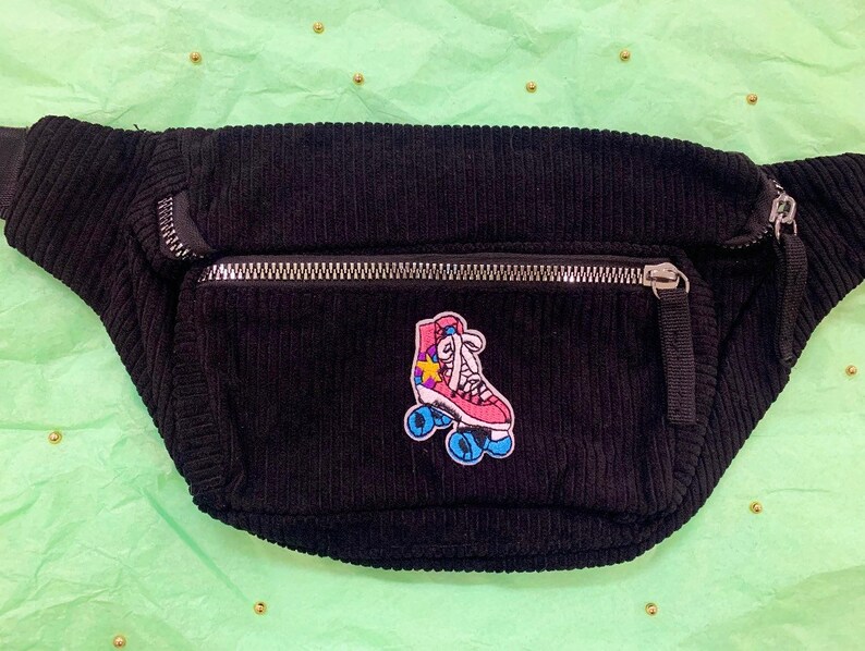 K&ouml;nnte beinhalten: Schwarze Cord-Bauchtasche mit einem rosa-blauen Rollschuh-Patch. Der Patch hat einen gelben Stern und wei&szlig;e Schn&uuml;rsenkel.