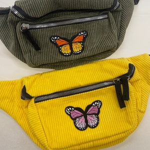 May include: Two fanny packs with butterfly patches. The top fanny pack is green corduroy with an orange butterfly patch. The bottom fanny pack is yellow corduroy with a pink butterfly patch.
