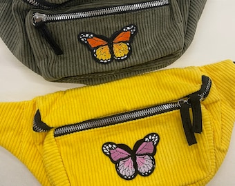 May include: Two fanny packs with butterfly patches. The top fanny pack is green corduroy with an orange butterfly patch. The bottom fanny pack is yellow corduroy with a pink butterfly patch.