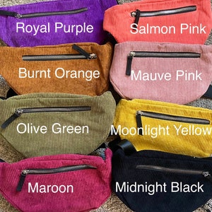May include: A collection of corduroy fanny packs in various colors, including Royal Purple, Salmon Pink, Burnt Orange, Mauve Pink, Olive Green, Moonlight Yellow, Maroon, and Midnight Black. Each pack features a zippered pocket and a black strap.