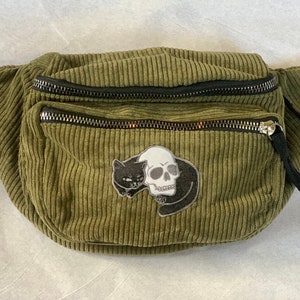 May include: Olive green corduroy fanny pack with black zippers and a black adjustable strap. The fanny pack features a patch with a black cat and a white skull design. The bag has two zippered compartments.
