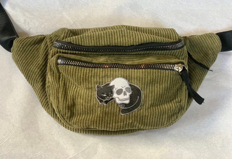 May include: Olive green corduroy fanny pack with black zippers and a black adjustable strap. The fanny pack features a patch with a black cat and a white skull design. The bag has two zippered compartments.