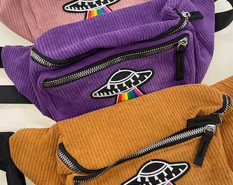May include: Three corduroy fanny packs in pink, purple, and brown. Each bag features a black and white UFO patch with a rainbow beam. The bags have black zippers and straps, offering a stylish and functional accessory.