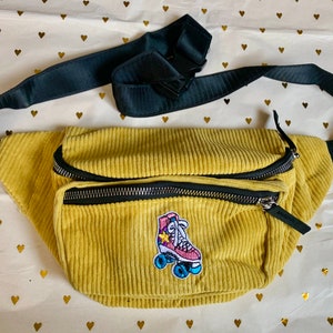 K&ouml;nnte beinhalten: Eine gelbe Cord-Bauchtasche mit einem schwarzen verstellbaren Riemen und einem bunten Rollschuh-Patch.
