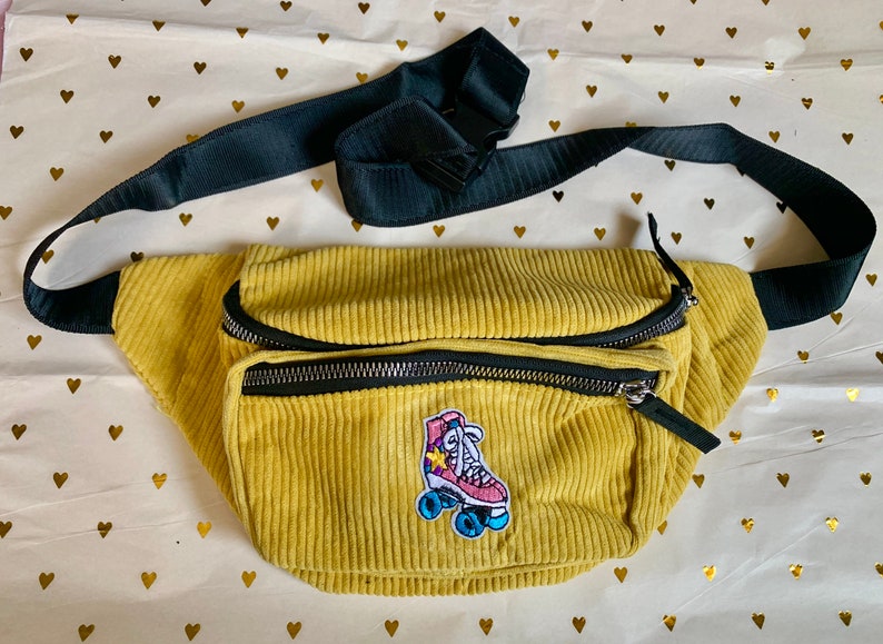 K&ouml;nnte beinhalten: Eine gelbe Cord-Bauchtasche mit einem schwarzen verstellbaren Riemen und einem bunten Rollschuh-Patch.