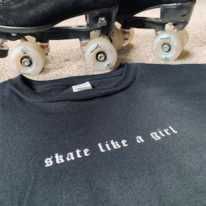 Puede incluir: Camiseta negra con texto blanco que dice "skate like a girl" en una fuente gótica.