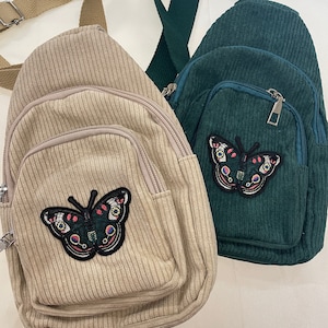 May include: Two corduroy crossbody bags with butterfly patches. One bag is beige and the other is green.