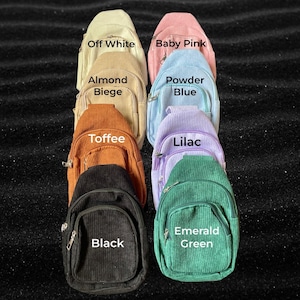 May include: Nine corduroy mini backpacks in a variety of colours, including off-white, baby pink, powder blue, lilac, toffee, black, and emerald green. Each backpack has a zip closure and a small pocket on the front. The backpacks are arranged in a semi-circle on a black surface.