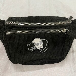 May include: Black corduroy fanny pack with two zippered compartments. A patch featuring a skull and a cat is sewn on the front. The adjustable strap is black. The bag is designed to be worn around the waist or across the body.