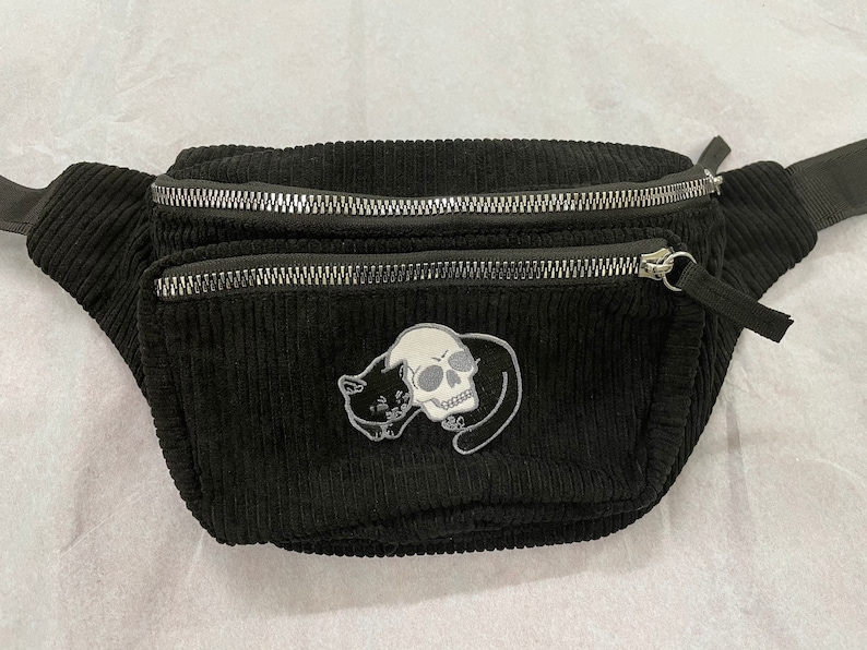 May include: Black corduroy fanny pack with two zippered compartments. A patch featuring a skull and a cat is sewn on the front. The adjustable strap is black. The bag is designed to be worn around the waist or across the body.