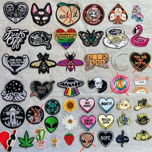 May include: A collection of embroidered iron-on patches featuring various designs, including a cat, a bee, a skull, a UFO, a sunflower, a heart, a moon, a flamingo, and a skull. The patches are colourful and have a variety of themes, including horror, space, and nature.