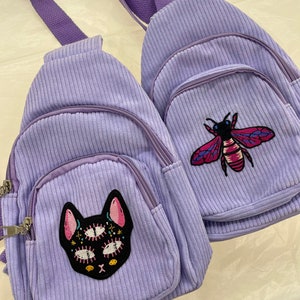 May include: Two small purple corduroy backpacks with embroidered patches. One backpack features a black cat with multiple eyes and the other has a bee with red and yellow accents.