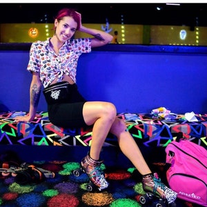 May include: A woman wearing a white and black patterned shirt, black shorts, and a black belt with a silver buckle is sitting on a blue bench. She is wearing roller skates with a leopard print design. A pink athletic bag is on the floor next to her.
