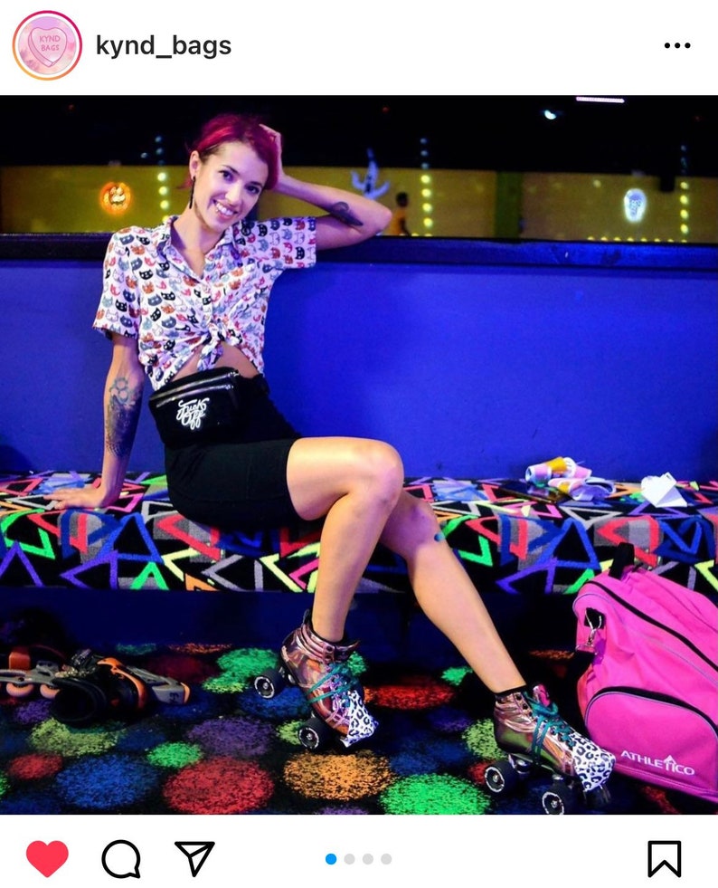May include: A woman wearing a white and black patterned shirt, black shorts, and a black belt with a silver buckle is sitting on a blue bench. She is wearing roller skates with a leopard print design. A pink athletic bag is on the floor next to her.