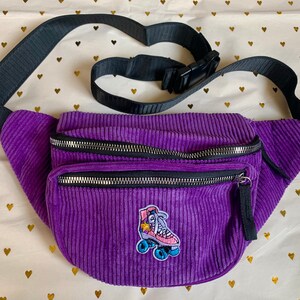 K&ouml;nnte beinhalten: Lilafarbene Cord-Bauchtasche mit einem verstellbaren schwarzen Gurt und einem Rei&szlig;verschluss. Die Bauchtasche hat einen Rollschuh-Aufn&auml;her mit einem Stern und einem Regenbogen.