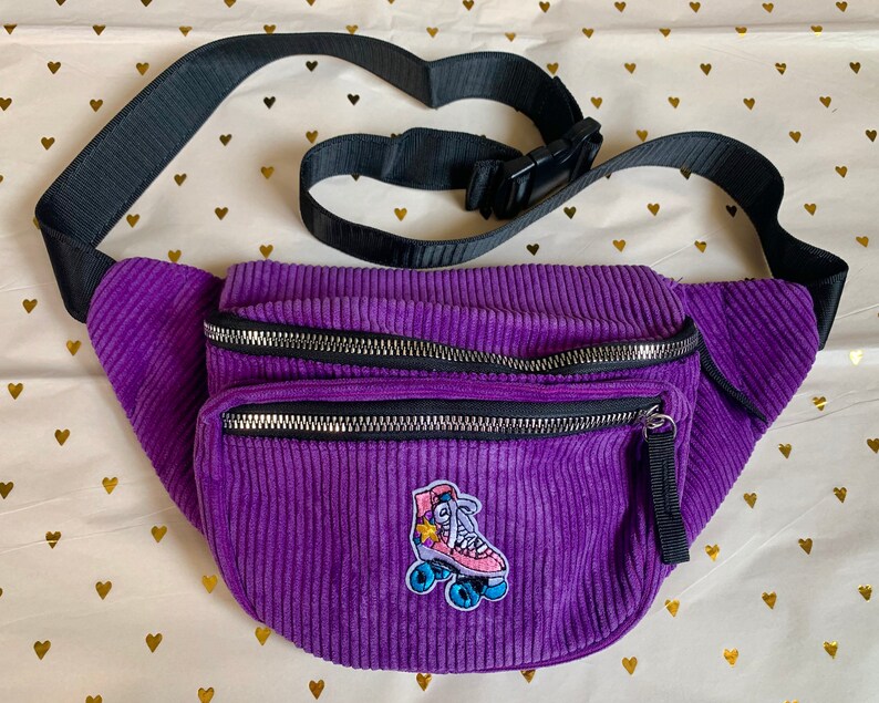 K&ouml;nnte beinhalten: Lilafarbene Cord-Bauchtasche mit einem verstellbaren schwarzen Gurt und einem Rei&szlig;verschluss. Die Bauchtasche hat einen Rollschuh-Aufn&auml;her mit einem Stern und einem Regenbogen.