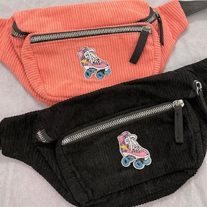 K&ouml;nnte beinhalten: Zwei G&uuml;rteltaschen aus Cord mit Rollschuh-Patches. Die obere G&uuml;rteltasche ist rosa und die untere G&uuml;rteltasche ist schwarz.