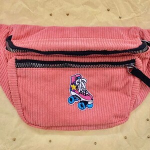 K&ouml;nnte beinhalten: Eine rosa Cord-Bauchtasche mit schwarzem Rei&szlig;verschluss und schwarzem Gurt. Die Bauchtasche hat einen gestickten Aufn&auml;her mit einer Rolle mit einem Stern.