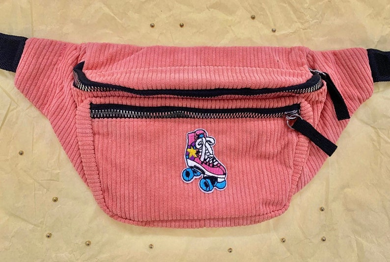 K&ouml;nnte beinhalten: Eine rosa Cord-Bauchtasche mit schwarzem Rei&szlig;verschluss und schwarzem Gurt. Die Bauchtasche hat einen gestickten Aufn&auml;her mit einer Rolle mit einem Stern.