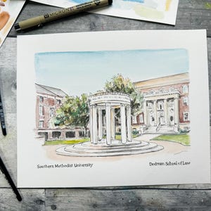 May include: Watercolor painting of the Southern Methodist University Dedman School of Law campus. The painting features a white, columned structure with a blue sky and green trees in the background. The text "Southern Methodist University" and "Dedman School of Law" are written below the image.