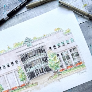 May include: Watercolor painting of Wayne State University Law School. The building is depicted in shades of gray with a glass facade, green trees, and red flowers. Three black pens are placed above the painting.