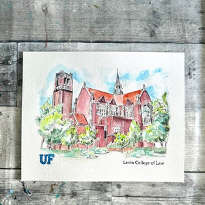 May include: Watercolor painting of the Levin College of Law building at the University of Florida. The building is red brick with a tall tower and a large, arched entrance. The painting is signed "UF" in blue and "Levin College of Law" in black.
