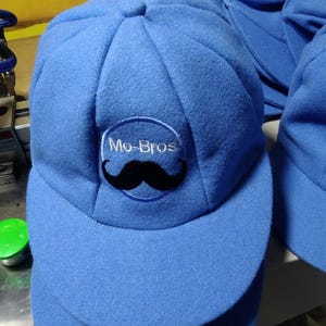 May include: A blue baseball cap with a curved brim. The cap features a circular patch with the text "Mo-Bros" and a black mustache graphic. The cap is made of a soft fabric and has a structured crown.