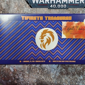 以下が含まれることがあります： 「Tifirets Treasures」の文字とライオンのロゴが入った製品パッケージ。パッケージは青と茶色の斜めストライプ模様です。Warhammer 40,000のロゴが上部にあります。赤い縁取りのカードのセットが右側にあります。