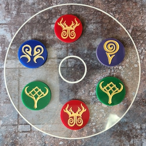 Könnte beinhalten: Ein klares, kreisförmiges Acrylteil mit weißem Rand, das sechs farbige, runde Token enthält. Die Token sind rot, blau, grün und lila, jedes mit einem goldfarbenen, stilisierten Design. Die Anordnung ist symmetrisch.