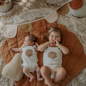 May include: Two babies, one wearing a white onesie with a seashell design and the other wearing a white onesie with a seashell design, lay on a brown and tan play mat with a fabric garland above them.
