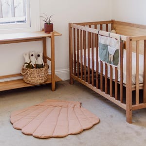 May include: A light brown quilted shell-shaped play mat for babies and toddlers. The mat is on a light brown carpet floor next to a wooden crib.