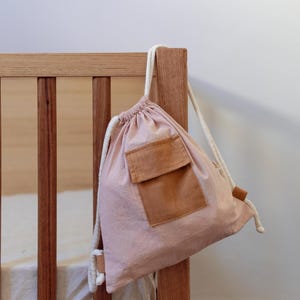 Peut inclure: Un sac à cordon rose avec une grande poche marron sur le devant. Le sac est suspendu à un lit bébé en bois.