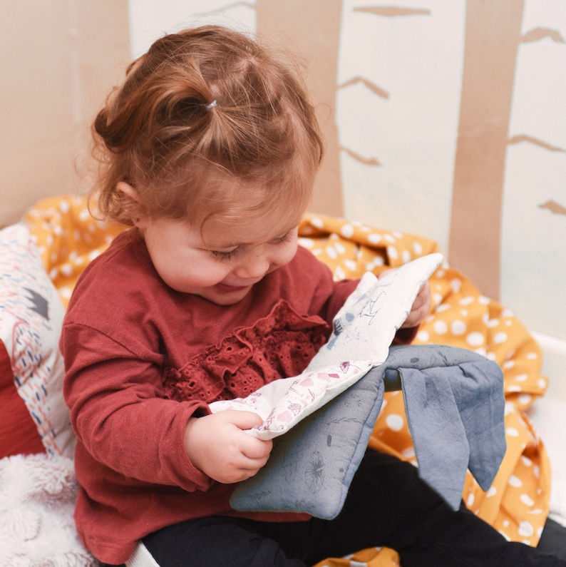 May include: A young child in a red and white dress is sitting on a yellow and white blanket. The child is holding a soft, gray and white stuffed animal with a floral pattern. The child is smiling and looking at the stuffed animal.