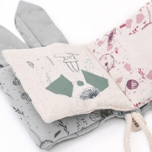 May include: A soft, fabric book for babies and toddlers with a woodland theme. The book features a green and white mushroom, a green forest, and a pink and white floral pattern. The book has two soft, gray bunny ears.