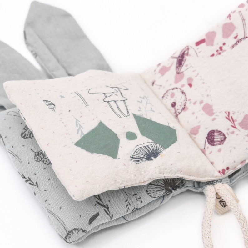 May include: A soft, fabric book for babies and toddlers with a woodland theme. The book features a green and white mushroom, a green forest, and a pink and white floral pattern. The book has two soft, gray bunny ears.