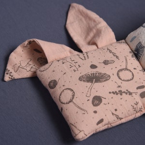May include: A pink fabric baby toy with a mushroom and leaf pattern. The toy is shaped like a bunny with two ears and a body.