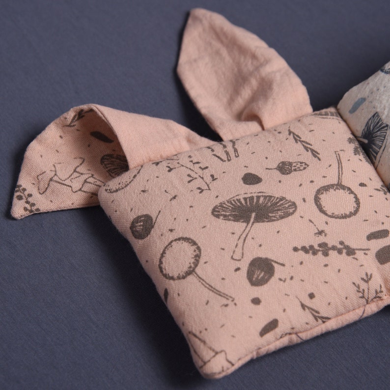 May include: A pink fabric baby toy with a mushroom and leaf pattern. The toy is shaped like a bunny with two ears and a body.