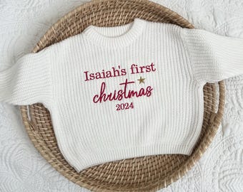 Op de afbeelding: Witte gebreide babytrui met de tekst "Isaiah's first christmas 2024" geborduurd in rood met een gouden ster.